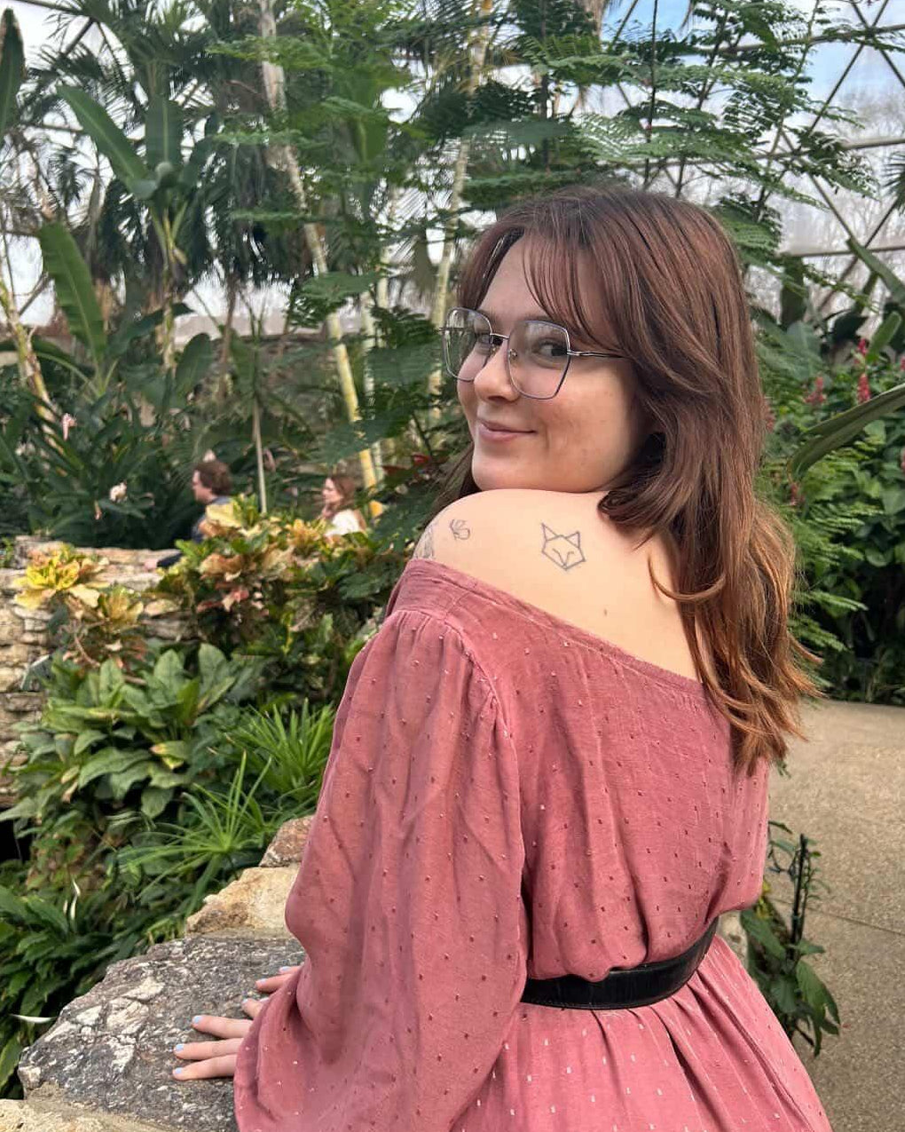 A woman with glasses and a small star tattoo on her shoulder sits on a stone ledge in a lush tropical greenhouse, wearing a dusty rose pink off-the-shoulder dress with a black waist belt.