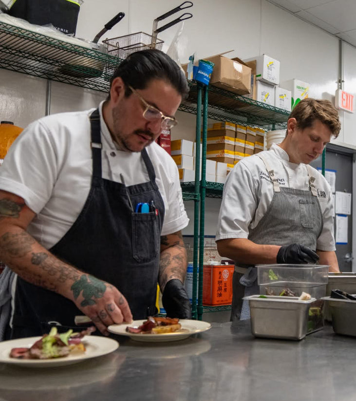 Two chefs in white shirts and aprons preparing food in a commercial kitchen, with one chef in the foreground plating a dish on a stainless steel counter.